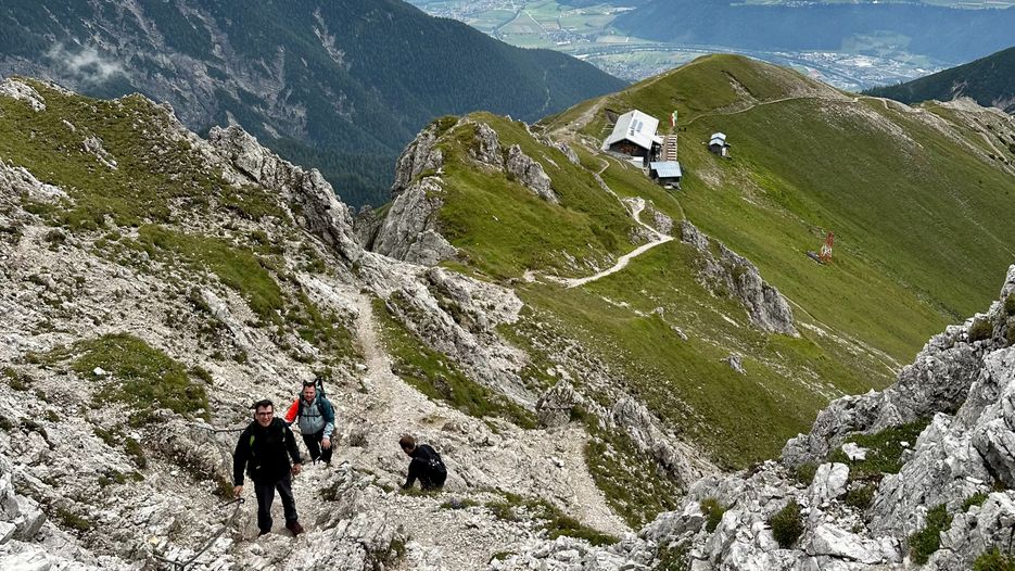 Berglandschaft mit Wanderern auf einem steilen Pfad, im Hintergrund eine Berghütte und grüne Wiesen.
