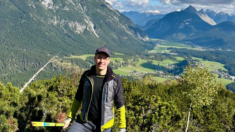 Ein Mann steht in der Natur mit Bergen im Hintergrund und einem Tal voller Bäume und Wiesen.