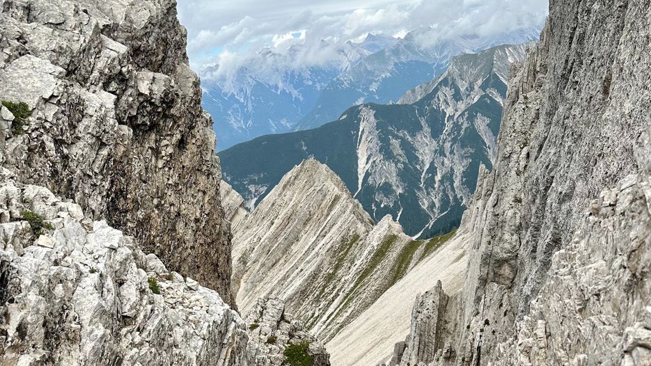 Berglandschaft mit schroffen Felsen und schmalen Tälern, umgeben von hohen Gipfeln und bewölktem Himmel.
