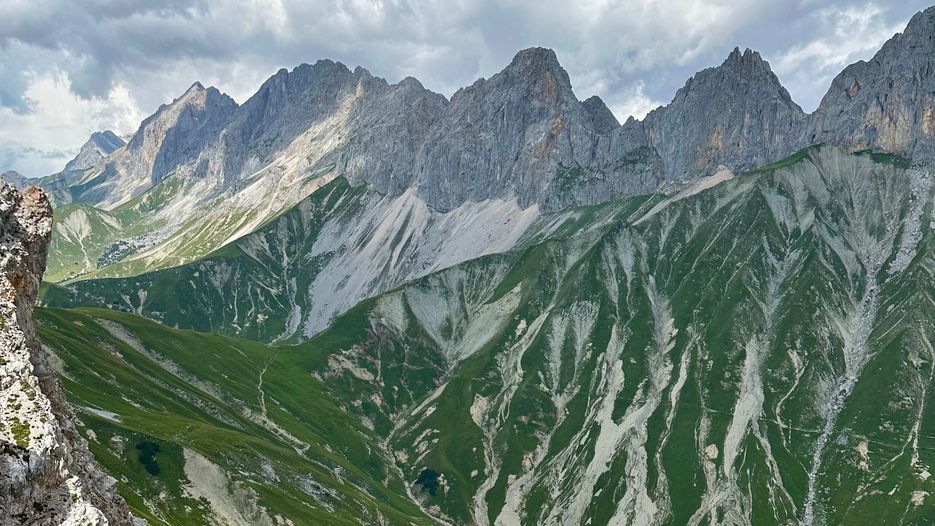 Berglandschaft mit steilen, grünen Hängen und schroffen Gipfeln unter einem bewölkten Himmel.
