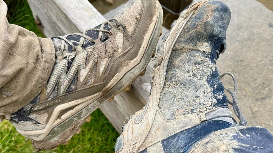 Zwei Paar schmutzige Wanderschuhe, die auf einer Holzbank liegen, umgeben von Gras und Wasser.