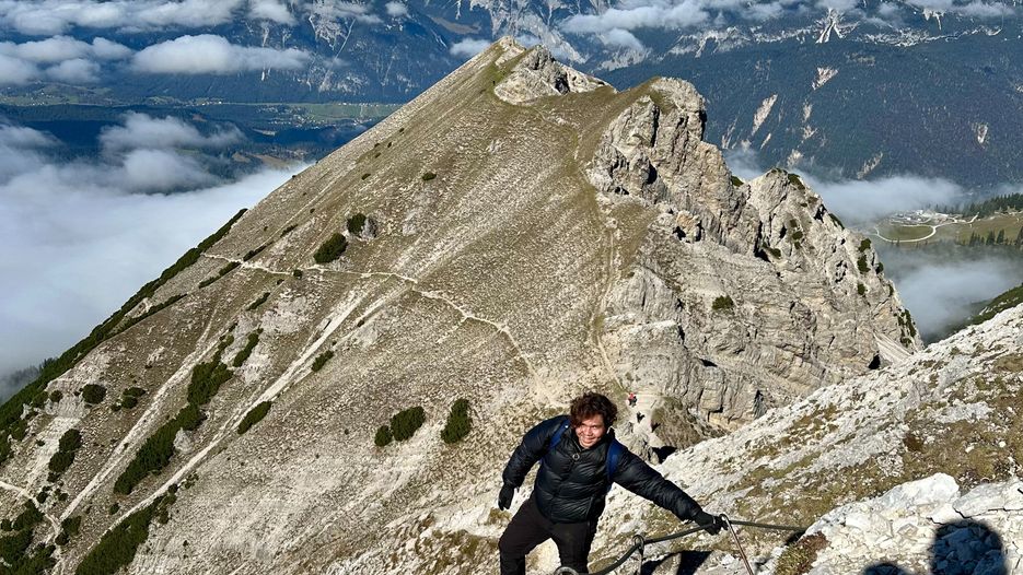 Eine Person klettert an einem steilen Berghang in den Dolomiten, umgeben von Wolken und Bergen.