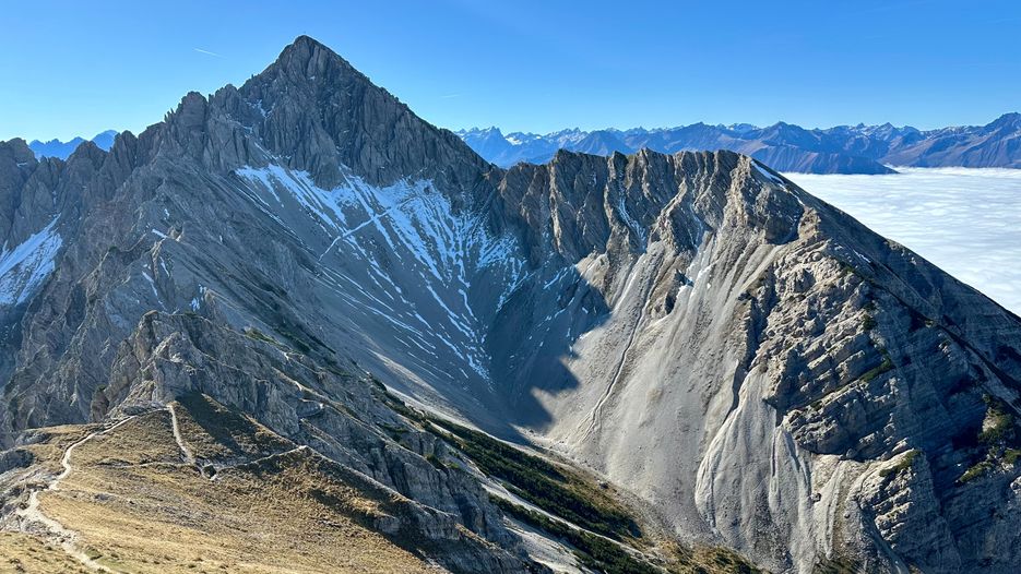 Eine beeindruckende Berglandschaft mit schroffen Gipfeln, schneebedeckten Bereichen und einem klaren blauen Himmel im Hintergrund.