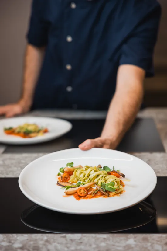 Chef serving a gourmet pasta dish with vegetables and tomato sauce on a white plate