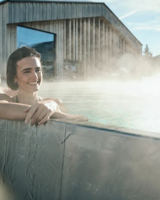 Lächelndes Paar entspannt im dampfenden Außenpool eines modernen Spa mit Holzgebäude im Hintergrund