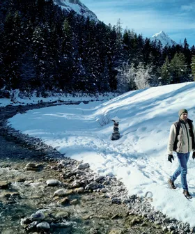 Two people walking along a snowy riverbank in a winter forest with mountain view