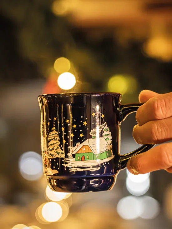 Hand holding festive Christmas mug with snowy village design and warm bokeh lights