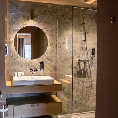 Modern hotel bathroom with stone walls, round mirror, walk-in shower and wooden vanity