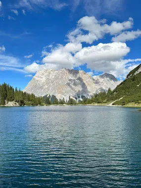 Klarer Bergsee mit Kiefernwald und felsigen Alpen-Gipfeln unter blauem Himmel mit Wolken