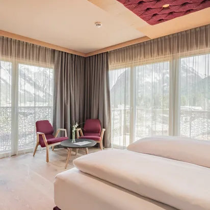 Modernes Hotelzimmer mit Doppelbett, Fernseher, Sitzecke und Bergblick durch große Fenster