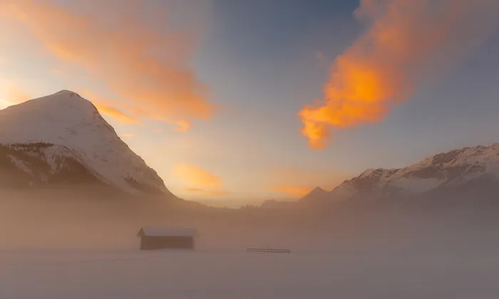 Snow-covered alpine landscape with wooden hut at sunset and glowing clouds