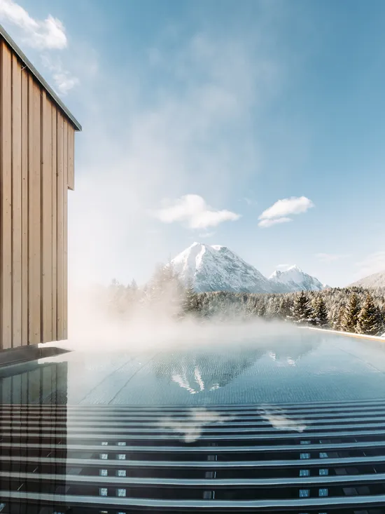 Dampf steigt aus Infinity-Pool mit Blick auf schneebedeckte Berge und Holz-Spa-Gebäude