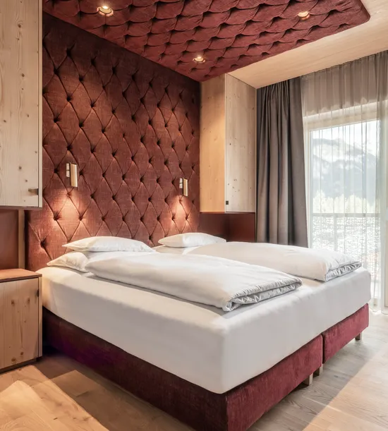 Modernes Hotelzimmer mit Doppelbett, bordeauxfarbener Polsterwand, Holzmöbeln und Bergblick