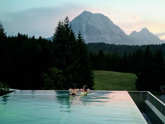 Couple relaxing in infinity pool overlooking forest and alpine mountains at sunset