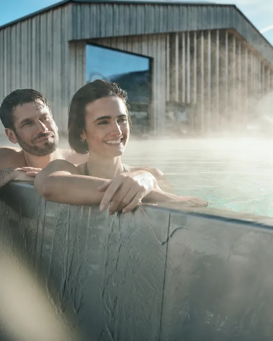 Lächelndes Paar entspannt im dampfenden Außenpool eines modernen Spa mit Holzgebäude im Hintergrund