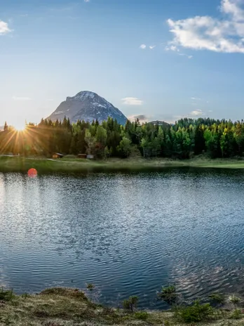 Mountain lake panorama with forest, calm water and sunset behind alpine peak