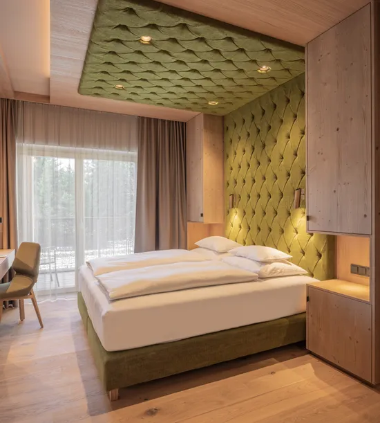 Modern hotel room with green upholstered bed and wood accents, natural light from balcony door