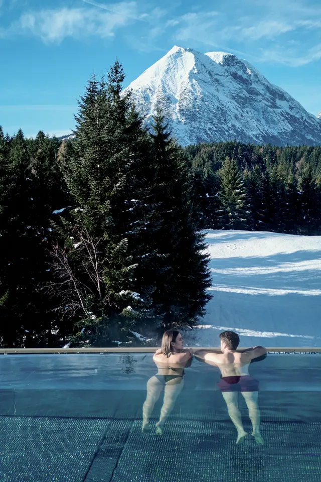 Couple relaxing in heated infinity pool overlooking snowy Alps and forest