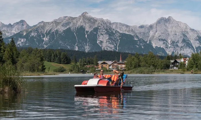 Touristen im Tretboot auf einem See mit Alpenpanorama im Hintergrund