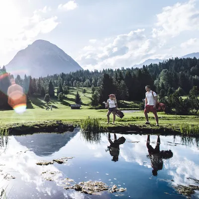 Golfers playing on a scenic mountain golf course with pond and pine trees
