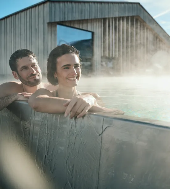 Lächelndes Paar entspannt im dampfenden Außenpool eines modernen Spa mit Holzgebäude im Hintergrund