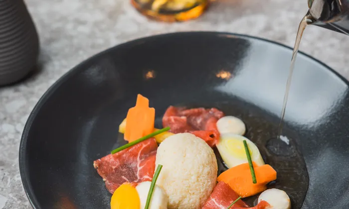Gourmet-Rindercarpaccio mit Gemüse und Brühe in schwarzer Schale auf Steintisch serviert