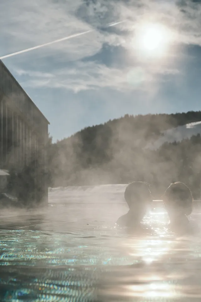 Paar entspannt in dampfendem Außenpool mit schneebedeckten Bergen und Sonne im Hintergrund