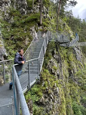 Man stands on metal walkway over rocky gorge in lush forested mountain area