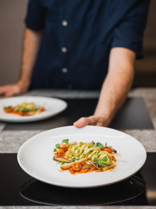 Chef serving a gourmet pasta dish with vegetables and tomato sauce on a white plate