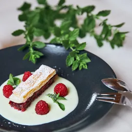 Himbeer-Mille-Feuille-Dessert mit Vanillesoße und Minze auf schwarzem Teller mit Latte im Hintergrund