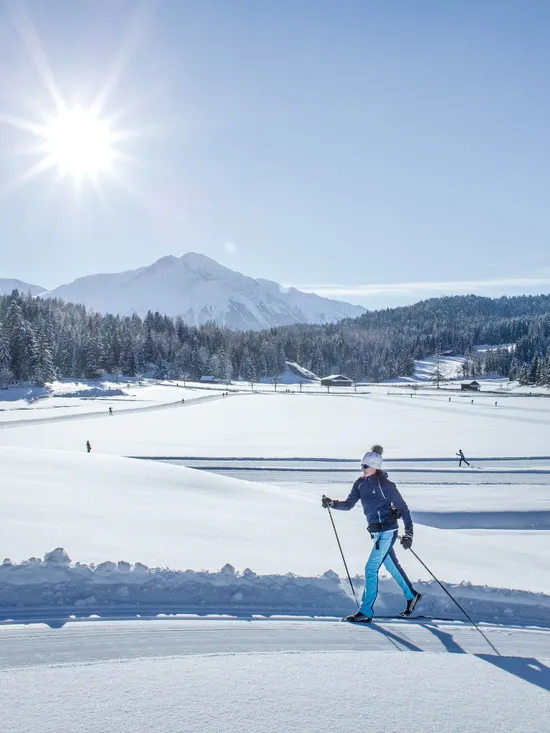 Woman cross-country skiing on snowy trail with alpine mountains and forest in sunlight