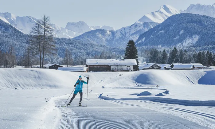 Cross-country skier on groomed snow trail with alpine village and snowy mountains in background