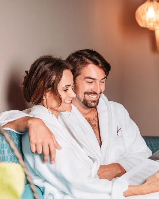 Smiling couple in spa robes reading brochure on wellness retreat sofa