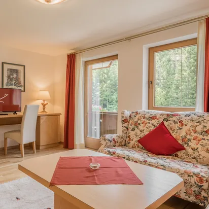 Cozy hotel room with floral sofa, wooden furniture, desk, TV, and large windows with red curtains