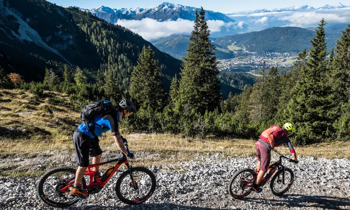 Mountainbiker auf steinigem Alpentrail mit Panoramablick auf schneebedeckte Gipfel und Waldtal