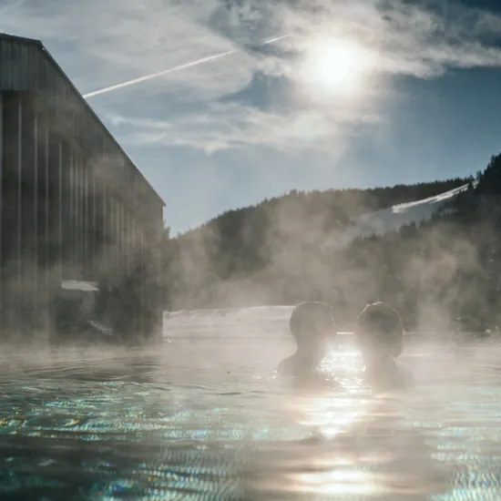 Paar entspannt in dampfendem Außenpool mit schneebedeckten Bergen und Sonne im Hintergrund