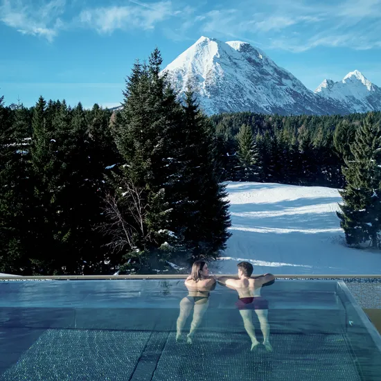 Couple relaxing in heated infinity pool overlooking snowy Alps and forest