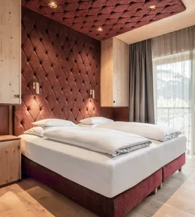 Modernes Hotelzimmer mit Doppelbett, bordeauxfarbener Polsterwand, Holzmöbeln und Bergblick