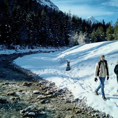 Zwei Personen gehen an einem verschneiten Flussufer im Winterwald mit Bergblick entlang