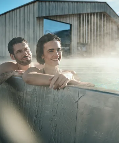 Lächelndes Paar entspannt im dampfenden Außenpool eines modernen Spa mit Holzgebäude im Hintergrund