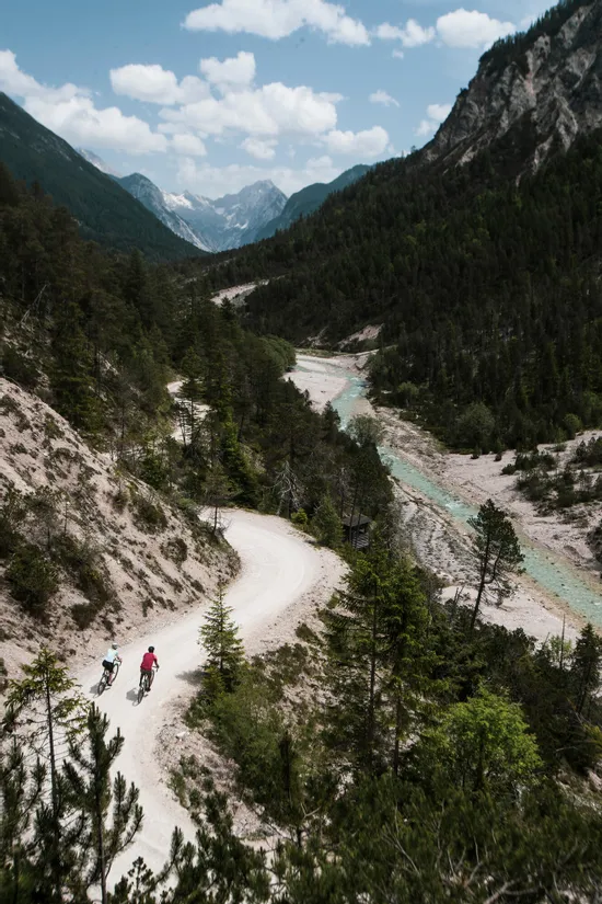Zwei Radfahrer auf einem kurvigen Bergpfad neben einem türkisfarbenen Fluss in den Alpen