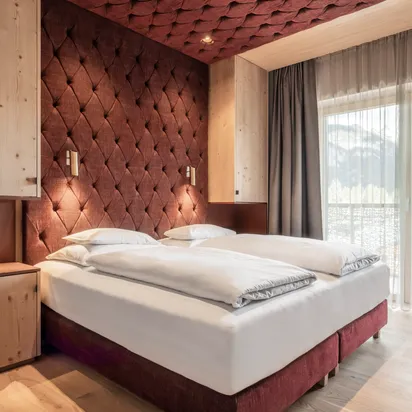 Modernes Hotelzimmer mit Doppelbett, bordeauxfarbener Polsterwand, Holzmöbeln und Bergblick