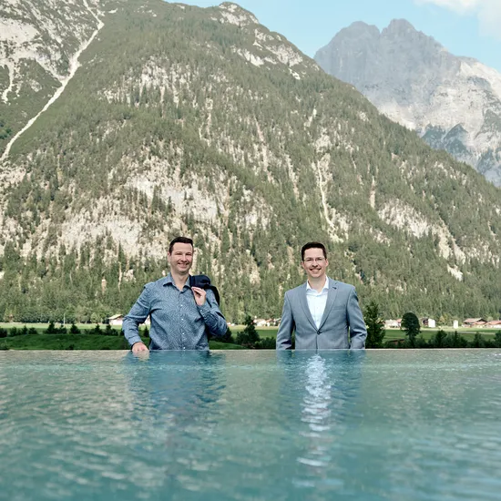 Zwei Männer in Businesskleidung stehen in einem Infinity-Pool vor alpiner Bergkulisse