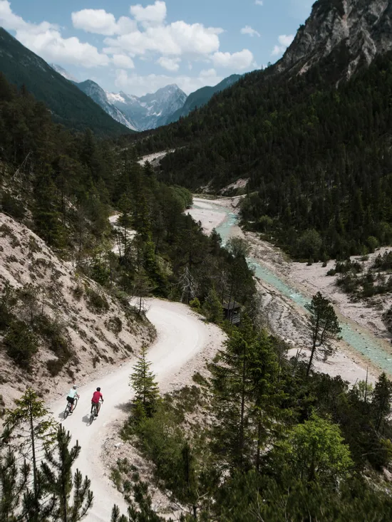 Zwei Radfahrer auf einem kurvigen Bergpfad neben einem türkisfarbenen Fluss in den Alpen