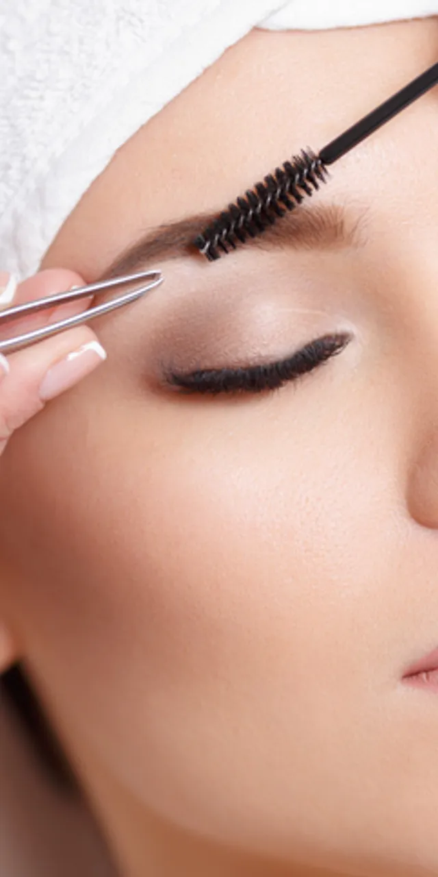 Woman receiving eyebrow shaping with tweezers and brush at beauty salon