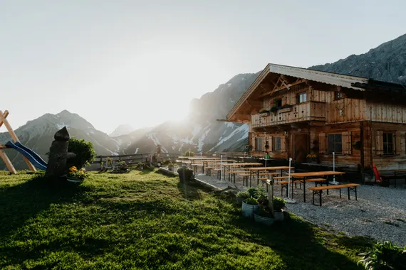 Rustic alpine hut with outdoor seating and mountain backdrop at sunset