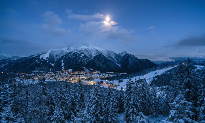 Verschneites Alpendorf mit beleuchteten Skipisten bei Nacht unter Vollmond