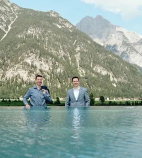 Zwei Männer in Businesskleidung stehen in einem Infinity-Pool vor alpiner Bergkulisse