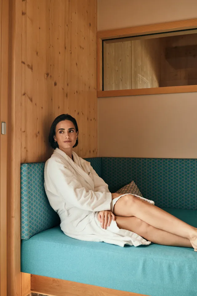 Woman relaxing on a turquoise bench in a spa lounge wearing a white bathrobe