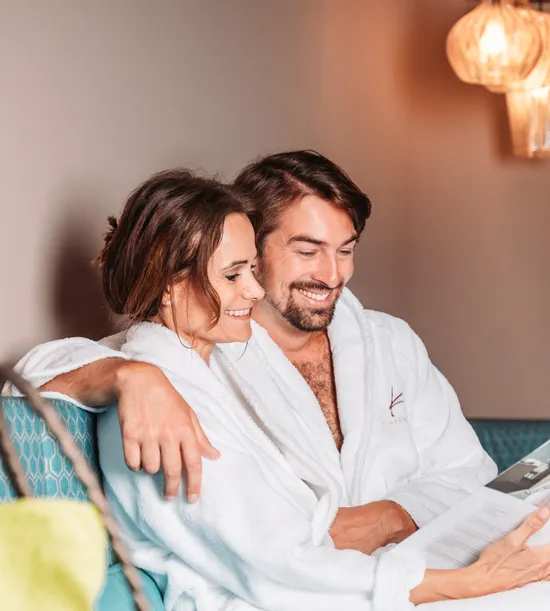 Smiling couple in spa robes reading brochure on wellness retreat sofa